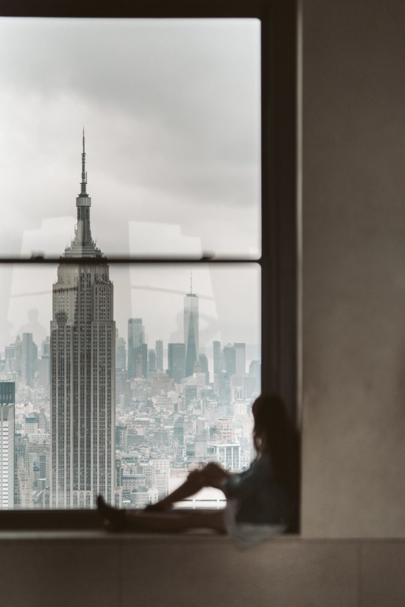 A woman sits in the window of an apartment and looks over the city alone.